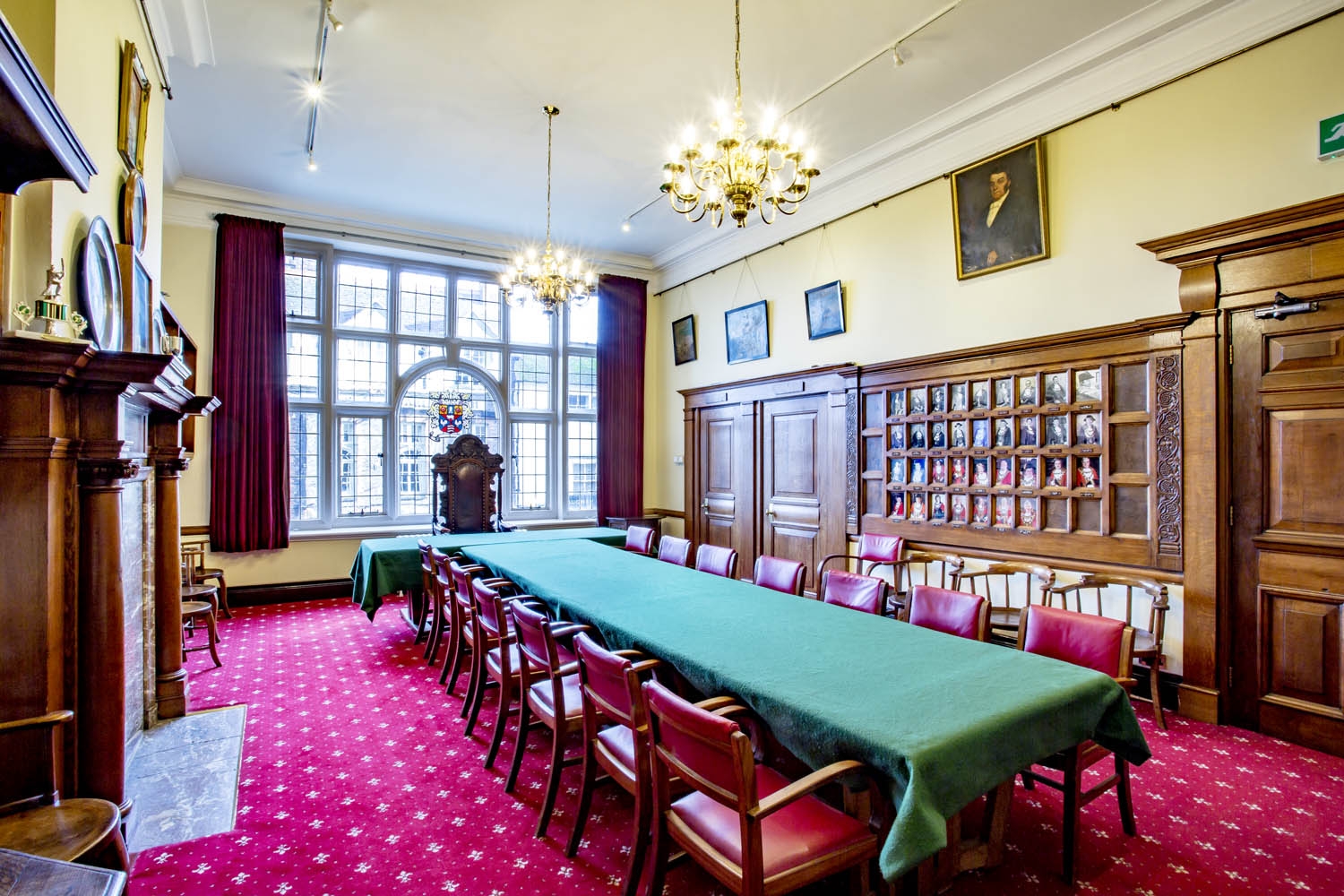 A grand, panelled room lined with portraits.  A very large window takes up one wall with a stained glass crest. A long table is lined by wooden and leather chairs