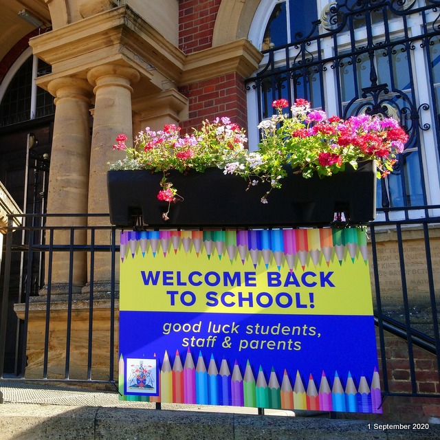 a bright and colourful sign wishing staff, students and parents good luck returning to school is attached to metal railings. There is a trough of flowers above and a brick and stone building behind