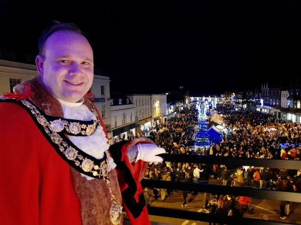 A man wearing a red ceremonial robe and chain of office smiles at the camera. He stands on a balcony and beyond is a street filled with people. There are decorative lights above a market