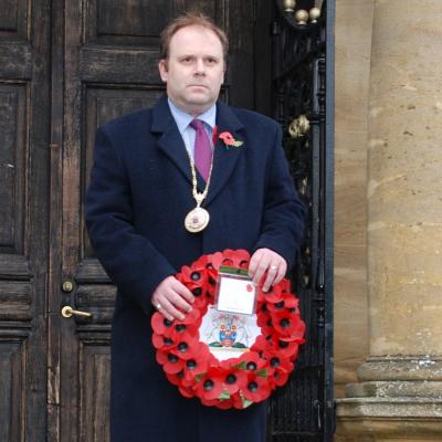 A smartly dressed man stands in front of a building.  He holds a wreath of poppies
