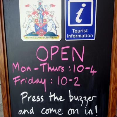 a black chalk board with a crest and a blue tourist information logo showing opening hours and the message press the buzzer and come on in