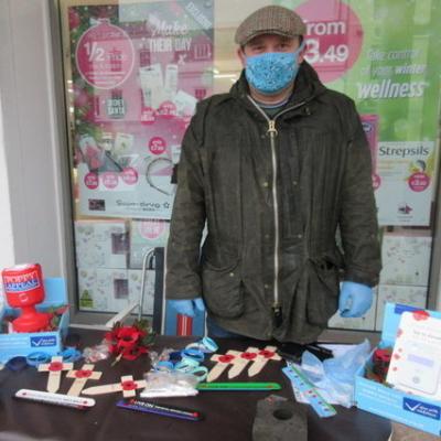 A man dressed for cold wet weather stands at a table selling poppies and collecting donations.  Click or tap to open the Mayor's Blog