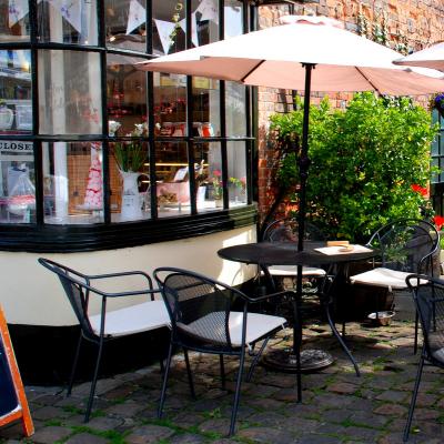 cafe tables and chairs with a parasol sit to the right of a building with a large window
