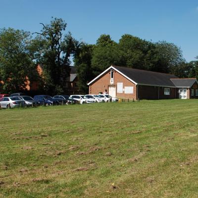 a field.  Behind it to the left is a car park full of cars, with a brick building to the right