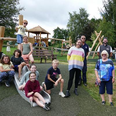 children are sitting on a slide, smiling to the camera.  Adults stand to the right. Behind them are wooden play structures