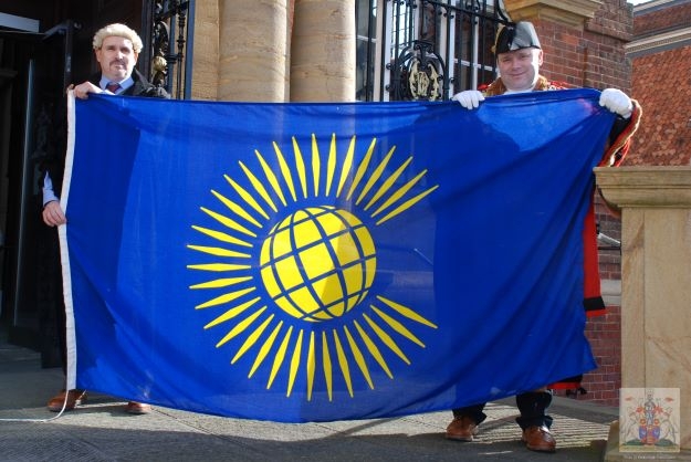 Photo of a woman and two men, all wearing gowns of office.  The man in the middle wears a mayoral chain and reads from a piece of paper