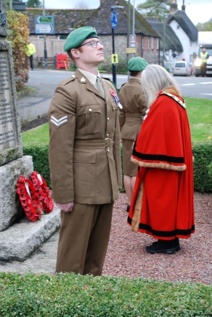 a cenotaph.  A poppy wreath is at the base.  Sideways on, two soldiers stand to attention while a woman in a red gown bows her head