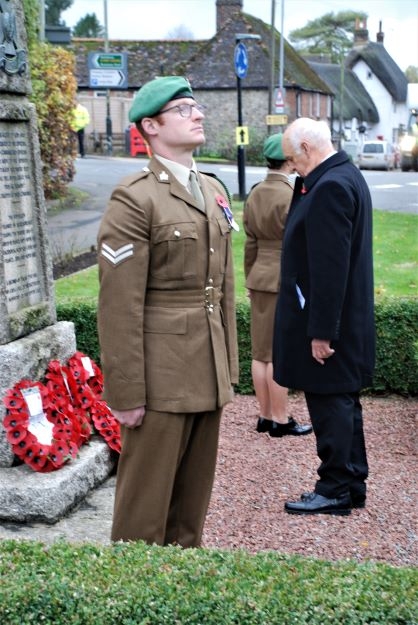 A man in a dark coat bows his head before a cenotaph, with a poppy wreath at the base.  Soldiers stand to attention at either side