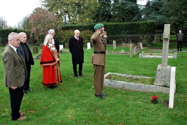 a cemetery.  On grass, a group of people face a grave.  Soldiers salute.  A poppy posy lies near the white headstone
