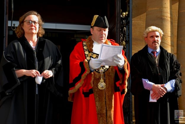 Photo of a woman and two men, all wearing gowns of office.  The man in the middle wears a mayoral chain and reads from a piece of paper