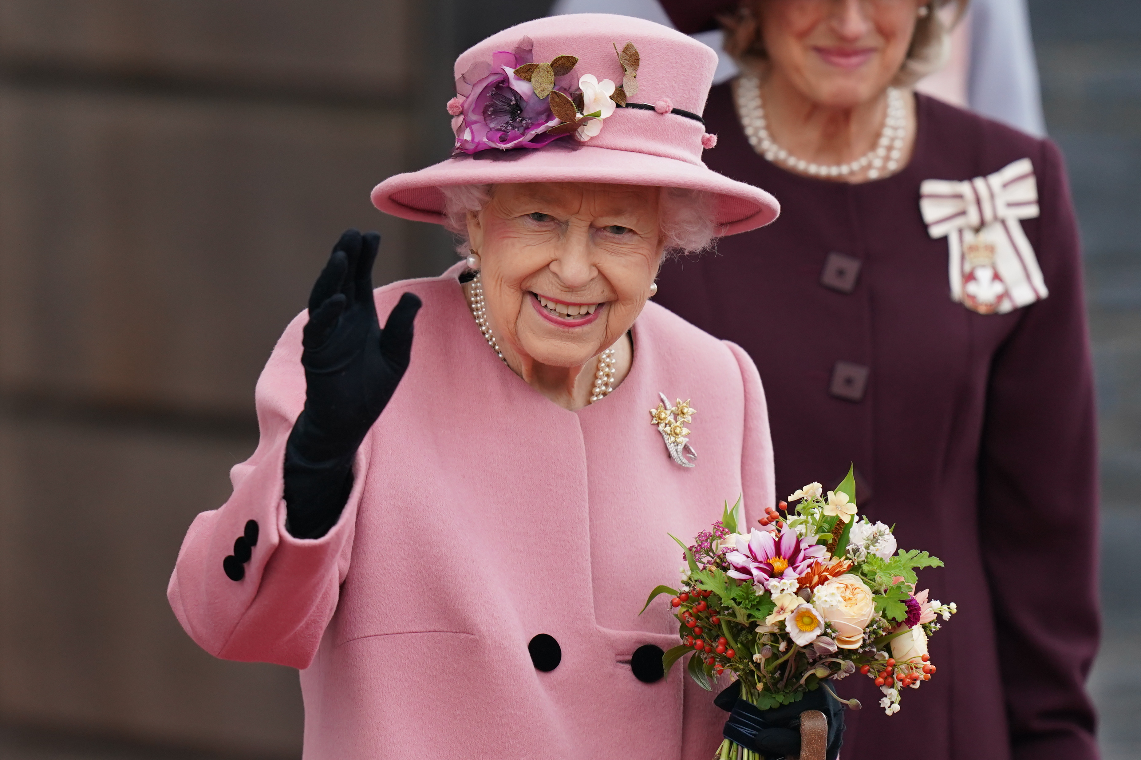 Photo of HM Queen Elizabeth II: an elderly, smiling woman waves. She wears matching pink coat and hat and carries a posy of flowers