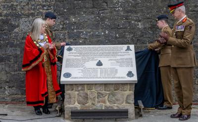on the left, a woman in red ceremonial robes and a soldier look to their right.  To the right, two men in army uniform look to the left.  Between them is a stone monument with an inscribed tablet on the top