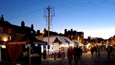 A street scene, with the sun setting.  There are market stalls in a row and shoppers in winter clothing