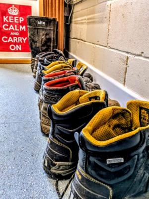 a row of boots lined up against a wall.  In the background a sign reads keep calm and carry on