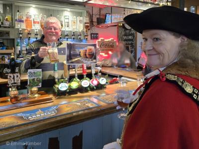 A woman in a red gown and black hat in front of a bar.  A smiling man behind the bar holds up a certificate