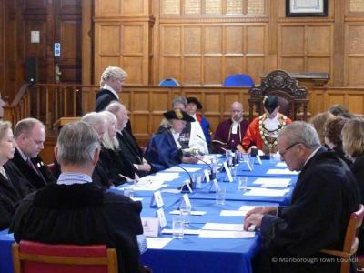 People in black robes are seated around a table.  A woman in a blue gown is writing, with a man standing behind her to witness