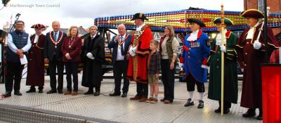people in ceremonial robes and chains stand on the platform of a fairground ride.  A man has a microphone in his hand