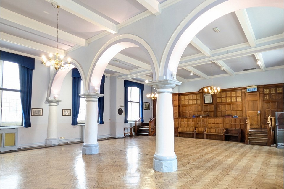 A large bright empty room with large windows along either side. Pillars and arches bisect part and there is a wood panelled platform at one end